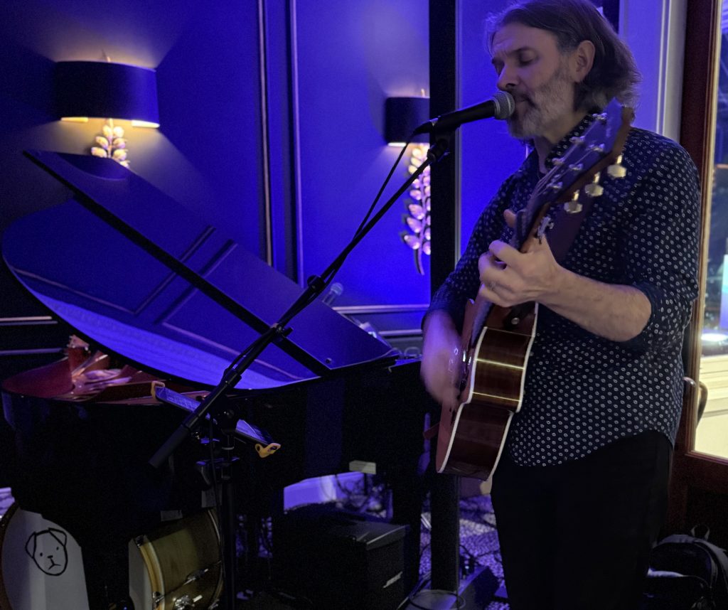a man playing guitar and singing into a microphone near baby grand with blue lighting