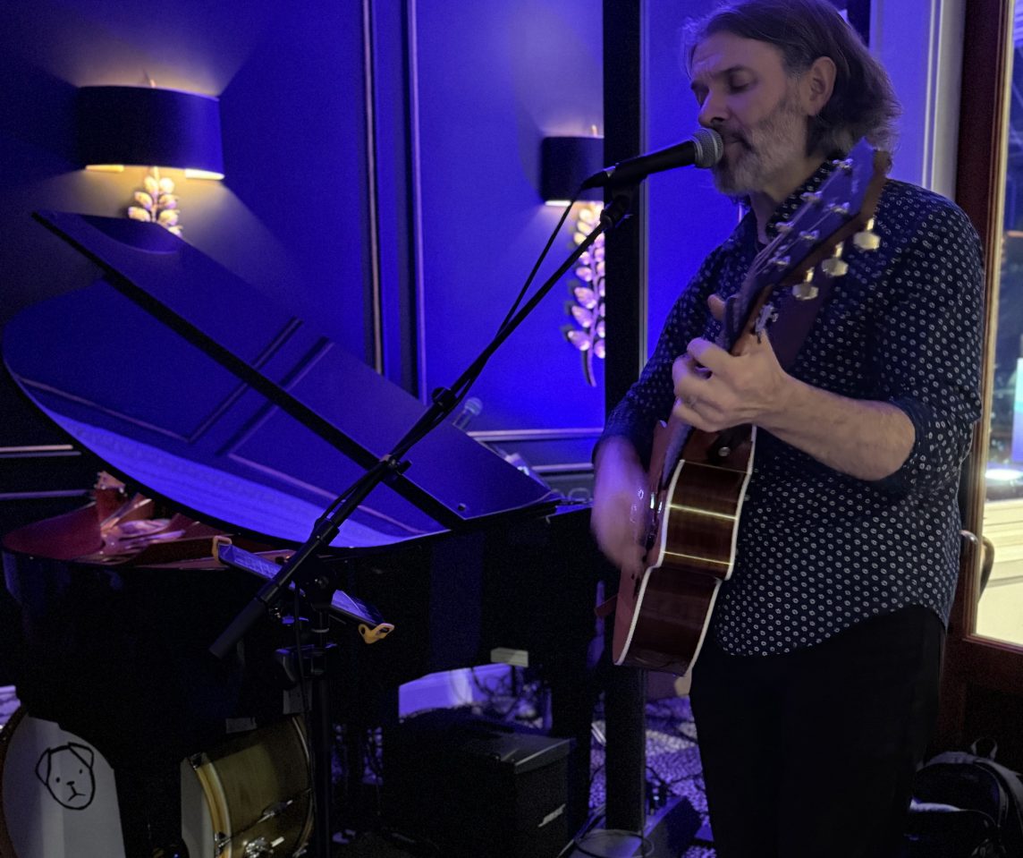 a man playing guitar and singing into a microphone near baby grand with blue lighting