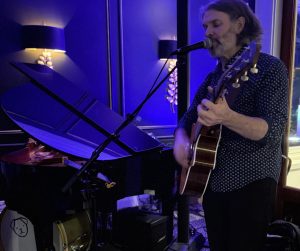 a man playing guitar and singing into a microphone near baby grand with blue lighting